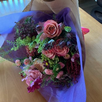 Flora Elements - Hand tied bouquet florist choice - A floral bouquet featuring pink roses, carnations, and various greenery, wrapped in purple and brown paper, placed on a wooden table. - Florists Near Me - Delafield, Wisconsin
