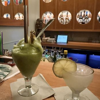 Daydream - Two cocktails are positioned on a wooden bar counter, each on a napkin. The left drink is a green frozen cocktail with a straw, a slice of lemon, and a spoon, while the right drink is a clear cocktail with a lemon slice garnish. The background features a wooden liquor display with circular cutouts showcasing various bottles, and a tablet device is visible on the bar shelf. - Cocktail Bars Near Me - Albuquerque, New Mexico