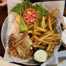 219 Burger - A metal tray lined with parchment paper holds a serving of French fries, a slice of tomato, lettuce, bacon strips, and a small cup of creamy dipping sauce. The tray is placed on a wooden table, with a person's arm and watch visible in the background.