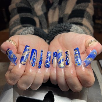 Gelanlis Nails - A person displays their hands with decorated nails featuring blue, gold, and white designs, along with rings and jewelry. - Nail Salons Near Me - Hammond, Indiana