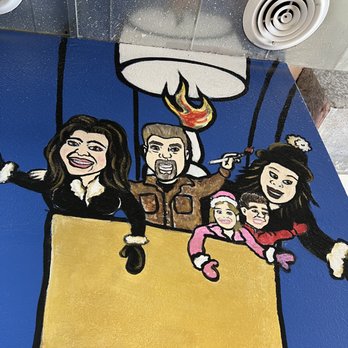 Sergio's Bakery and Cafe - The image shows a cartoon-style mural of a family of five in a hot air balloon, painted on a wall with a blue background. The family includes two adults and three children, all smiling and dressed in colorful clothing. The man holds a torch with a flame, and the balloon is depicted with a large yellow basket. The mural is painted with bold black outlines and vibrant colors, creating a playful and lively scene. - Bakeries Near Me - Albuquerque, New Mexico