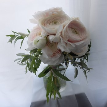 Tiny Flower - A bridal bouquet of pale pink ranunculus and trailing willow leaves. - A bouquet of pale pink ranunculus flowers with multiple layers of delicate petals, accompanied by green foliage, arranged in a clear glass vase. The background is light and softly lit, highlighting the soft colors and textures of the flowers and leaves. - Florists Near Me - Toronto, Ontario