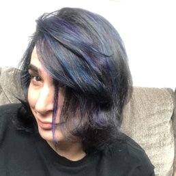 A woman with shoulder-length, layered hair styled with a side parting, featuring shades of blue and purple highlights. She is sitting on a brown textured couch with a neutral background, wearing a black top, and looking slightly to the side with a subtle smile.