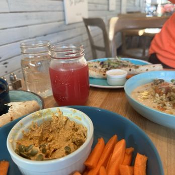 Just BE Kitchen - AMAZING DRINKS!!! - The image shows a wooden table with multiple dishes including hummus with pumpkin seeds, sliced carrots, naan bread, a bowl of soup, and a pink beverage in a jar. A person wearing an orange shirt is partially visible on the right side of the image. The background features a wall with horizontal wooden planks and some chairs, indicating an indoor or patio dining area at Just BE Kitchen. - Gluten-Free Near Me - Boulder, Colorado