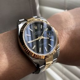 A close-up of a Rolex Oyster Perpetual Datejust wristwatch featuring a blue dial, gold fluted bezel, and a two-tone bracelet with stainless steel and gold. The watch displays the time with gold hands and hour markers, and includes a date window with a magnifying cyclops lens. The wristwatch is worn on a person's wrist, with a background of window blinds and a neutral-colored surface.