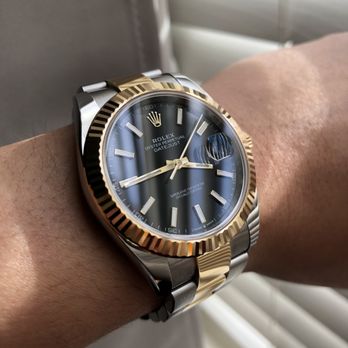 Rolex Boutique - A close-up of a Rolex Oyster Perpetual Datejust wristwatch featuring a blue dial, gold fluted bezel, and a two-tone bracelet with stainless steel and gold. The watch displays the time with gold hands and hour markers, and includes a date window with a magnifying cyclops lens. The wristwatch is worn on a person's wrist, with a background of window blinds and a neutral-colored surface. - Watches Near Me - Boston, Massachusetts