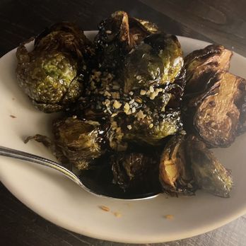 Rooftop - Brussels sprouts $12.00 - Charred Brussels sprouts garnished with chopped nuts are presented on a white dish, with a spoon on the side, likely served at a restaurant. - New American Near Me - Walnut Creek, California