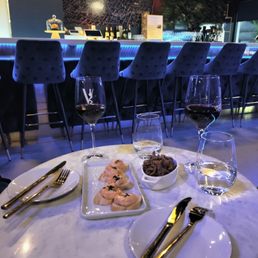 Garlic shrimp - A marble-topped round table in a restaurant setting is arranged with two glasses of red wine, a small water glass, and a plate with appetizers, possibly seafood or chicken. Silverware is placed on small white plates on either side of the table. The background shows a bar with a blue-lit counter, high-backed upholstered chairs, and a dark, reflective wall with a textured pattern. The lighting is low, creating a cozy atmosphere.