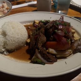 A restaurant plate contains beef stir-fry with sliced onions, peppers, and herbs, accompanied by a mound of white rice. The dish is served with a sauce and garnished with chopped herbs. The setting includes a wooden table with glasses and other dishes visible in the background.
