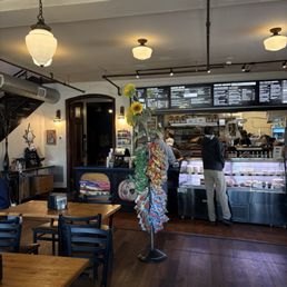 Inside of the restaurant - A cafe or bakery interior featuring a display case with baked goods, seating with wooden tables and chairs, and a sunflower decoration in the center.