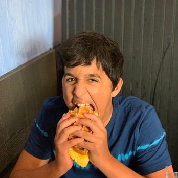 D’ Lua Pancake & Burrito House - Best waffle egg sandwich - A boy in a blue shirt is biting into a large sandwich or burger at a restaurant, with a dark wooden wall behind him. - Mexican Near Me - Moline, Illinois