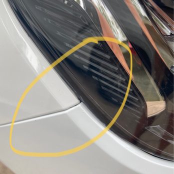 Caliber Collision - The image features a close-up of a vehicle's headlight, which has a visible crack highlighted by a yellow circle. The car's body is white, and the headlight has a dark tint with reflective surfaces. The background shows reflections of trees and surroundings on the headlight's surface. - Body Shops Near Me - Katy, Texas