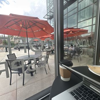 Cafe Select - Outside seating area - The image captures a view from inside a cafe looking out through a large glass window. Outside, there are several tables with black chairs and large red umbrellas providing shade. Some customers are seated at the tables, and the scene includes a street with parked cars and pedestrians. Inside the cafe, part of a laptop, a disposable coffee cup, and a plate are visible on a black table. The building's glass exterior reflects the sky and surrounding structures. - Coffee & Tea Near Me - Fort Lee, New Jersey