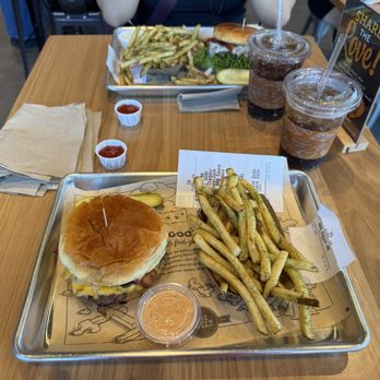 The Melt - A metal tray holds a cheeseburger with a bun, cheese, and a toothpick, alongside seasoned fries and a small container of dipping sauce. Two iced drinks with straws are on the table, along with a paper receipt and a napkin. The setting appears to be a casual restaurant with a wooden table and other patrons visible. - Burgers Near Me - Tempe, Arizona