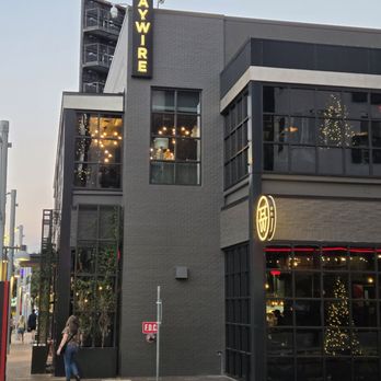 Haywire - The image shows a modern, multi-story building with large glass windows and exterior signage. The sign for Haywire is vertically lit in yellow, and a circular sign with a Chinese character is also visible. Interior lighting and Christmas decorations can be seen through the windows. The scene includes a sidewalk with a woman walking and a street with a red curb, indicating an urban setting. - New American Near Me - Austin, Texas