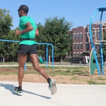 Park 578 - A person wearing sunglasses, a turquoise shirt, and black shorts is running in a park with playground equipment and trees. - Parks Near Me - Chicago, Illinois