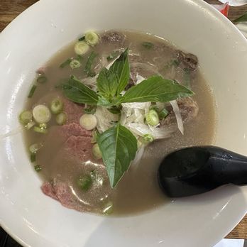 Link Thai - Beef Pho'' (Beef Noodle Soup) - A bowl of beef pho with broth, sliced beef, green onions, and fresh basil leaves, served with a black spoon. - Thai Near Me - Brooklyn, New York