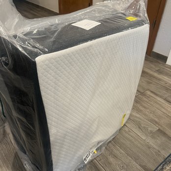 Nellis Auction - A mattress with a quilted white surface is wrapped in clear plastic and leaning against a wall in a room with wood flooring. The room has a wooden door and a gray wall, with some visible furniture and fixtures in the background. - Auction Houses Near Me - Mesa, Arizona