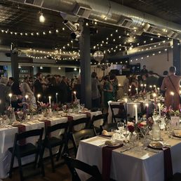 A large banquet hall features long tables with white tablecloths, red napkins, and floral centerpieces illuminated by candlelight. Guests are dressed in formal attire, mingling and socializing. The ceiling is decorated with string lights and a disco ball, creating a festive atmosphere. The background includes a bar area and a DJ setup, indicating a celebratory event.