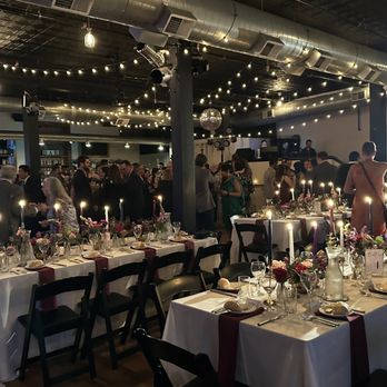 Floral Glory Design - A large banquet hall features long tables with white tablecloths, red napkins, and floral centerpieces illuminated by candlelight. Guests are dressed in formal attire, mingling and socializing. The ceiling is decorated with string lights and a disco ball, creating a festive atmosphere. The background includes a bar area and a DJ setup, indicating a celebratory event. - Floral Designers Near Me - Naperville, Illinois