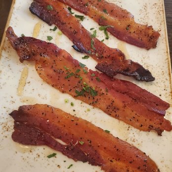 First Watch - Million dollar bacon looks like about $999,999 short - Four strips of cooked bacon with a crispy texture and visible seasoning, garnished with chopped green herbs, are arranged on a rectangular ceramic plate. The bacon appears to be seasoned with black pepper and possibly other spices, and the plate has a light speckled design with some seasoning or spice residue around the edges. - Breakfast & Brunch Near Me - Centennial, Colorado