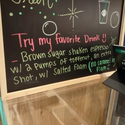 a menu on a chalkboard in a coffee shop - The image shows a chalkboard sign with handwritten text in pink, green, and white, promoting a Starbucks drink called 'Sumatra.' The sign encourages customers to try the drink and describes its ingredients, including brown sugar, shaken espresso, and salted foam, with a note about no caramel in the foam. A small digital menu display is positioned to the right, showing the drink name and category, with a wooden frame around the chalkboard.