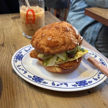 Woon - Pork Chop Sandwhich - A fried chicken sandwich with pickles and lettuce on a decorative plate, accompanied by iced coffee in a glass with a red logo. - Chinese Near Me - Pasadena, California