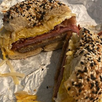 Time 4 Bagels - Bodega Bagel - A bagel sandwich with a sesame seed topping, filled with melted cheese, ham, and sausage, cut in half to reveal the ingredients. The sandwich is placed on crumpled foil, with some cheese and crumbs scattered around. The bagel appears toasted and the ingredients are layered inside. - Bagels Near Me - Gainesville, Virginia