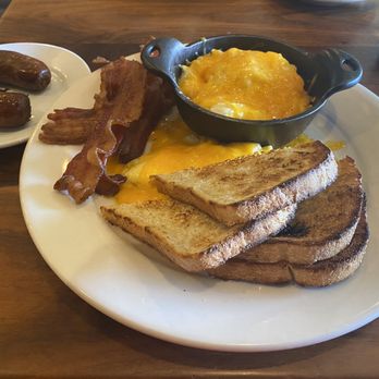 Bella's Cafe - Two Farm Fresh Eggs-Any style with a side of cheese grits, bacon and sausage. - A breakfast plate featuring scrambled eggs with cheese, toasted bread, bacon, sausage links, and a small skillet of cheese grits, served on a white plate. - Breakfast & Brunch Near Me - New Haven, Connecticut