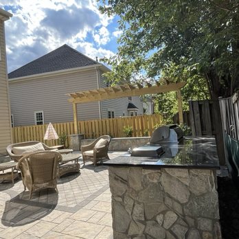 NOVA Landscape Construction - Island with built-in grill and cooktop. Paver patio. seating wall with trellis. - Residential backyard patio featuring outdoor seating, a built-in grill, and a pergola, enclosed by a wooden fence and surrounded by trees. - Snow Removal Near Me - Chantilly, Virginia