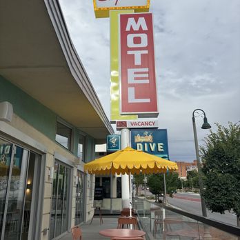 Vegan Vato - This is the location of where you can find really good vegan.  The 3 amigos are very good.  It is hard to find good vegan cheese. - Outdoor patio with pink chairs and a yellow umbrella in front of a restaurant. The restaurant has large glass windows and doors, with signage for the Imperial Motel and Dive. The scene includes a sidewalk, street, and trees, with a cloudy sky overhead. - Food Trucks Near Me - Albuquerque, New Mexico