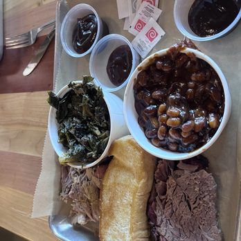 City Barbeque - Two Meats, Two Sides - The image shows a metal tray containing barbecue food items such as sliced beef, a fried bread roll, cooked collard greens, and baked beans in sauce. Two small cups of dark soda and condiment packets are also on the tray. The tray is placed on a wooden table, with a fork and knife visible nearby. The setting appears to be a casual dining environment. - Barbeque Near Me - Lafayette, Indiana