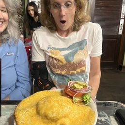 Volcano burger - A woman with glasses and curly, shoulder-length hair, wearing a graphic t-shirt, is seated at a table in a restaurant, reacting with surprise at a large cheeseburger covered in melted cheese. The burger has toppings including pickles, onions, and lettuce, and is served on a plate. The background shows framed photos on the wall and a wooden door, indicating a casual dining setting.