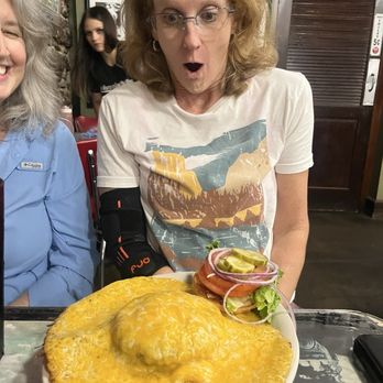 Ozark Cafe - Volcano burger - A woman with glasses and curly, shoulder-length hair, wearing a graphic t-shirt, is seated at a table in a restaurant, reacting with surprise at a large cheeseburger covered in melted cheese. The burger has toppings including pickles, onions, and lettuce, and is served on a plate. The background shows framed photos on the wall and a wooden door, indicating a casual dining setting. - American Near Me - Jasper, Arkansas