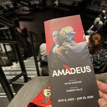 Steppenwolf Theater - A person holds a theater ticket for the production 'Amadeus' at Steppenwolf Theater, featuring images of characters from the play. The ticket displays the show dates from November 6, 2025, to January 25, 2026, and includes the names of the writer and director. The background shows the theater's audience seating area with several attendees seated and waiting for the performance. - Performing Arts Near Me - Chicago, Illinois