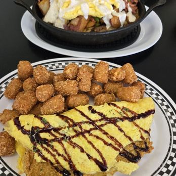 Lighthouse Diner - FGT Omelette - A breakfast meal includes an omelette topped with sauce and garnished with herbs, accompanied by fried chicken bites on a checkered plate. In the background, a skillet contains breakfast hash with eggs, vegetables, and meats, served on a white oval plate. The setting appears to be a diner or casual restaurant. - American Near Me - Jupiter, Florida