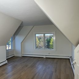 Custom windows - The image depicts an empty attic room with sloped ceilings on both sides, a large central window providing a view of trees outside, and hardwood flooring. The walls and ceiling are painted white, and baseboard heaters are visible along the lower walls. The space appears well-lit with natural light from the window.