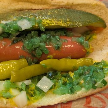 Devil Dawgs -  South State - Mmmm. - A close-up image of a hot dog in a bun, topped with sliced pickles, chopped onions, relish, and mustard. The hot dog is placed on a piece of paper, showing the toppings clearly. The bun appears soft and slightly toasted. - Hot Dogs Near Me - Chicago, Illinois