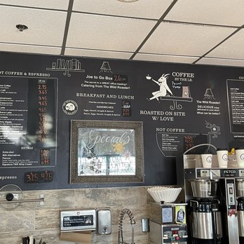 The Wild Roaster - A black chalkboard menu at The Wild Roaster coffee shop features sections for hot coffee, espresso, breakfast, lunch, and cold drinks. It includes prices for various beverages such as drip brew, cappuccino, latte, and cold drinks like iced coffee and lattes. The menu also highlights the shop's roasted-on-site coffee and catering options. The ceiling has a grid pattern with ceiling tiles, and part of the coffee station with equipment and supplies is visible below the menu. - Coffee & Tea Near Me - Deerfield, Illinois