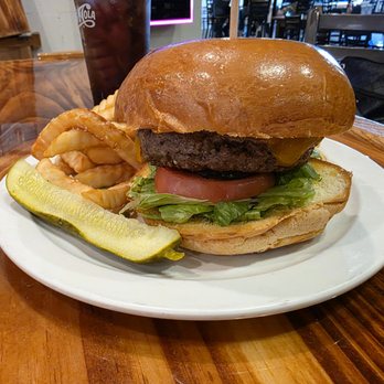 Bearded Monkey - Classic Burger - A burger featuring a beef patty, lettuce, tomato, and cheese on a toasted bun is served on a white plate. The plate also includes a pickle spear, curly fries, and a glass of soda in the background. The setting appears to be a casual dining restaurant with wooden tables and a lively atmosphere. - Chicken Wings Near Me - Fredericksburg, Virginia