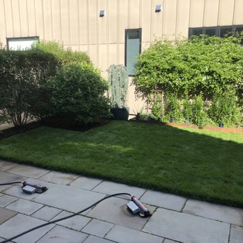 Lawn Jon Services - A backyard garden features a well-maintained lawn, bushes, potted plants, and a building with windows in the background. Gardening tools are on the patio. - Landscaping Near Me