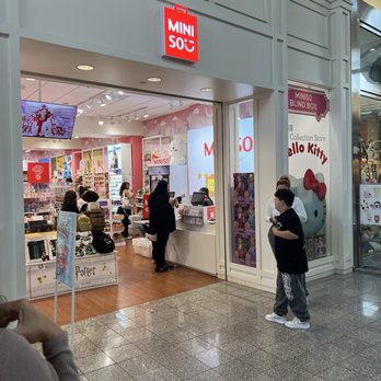 Miniso - The image shows the storefront of Miniso in Temecula, CA, with a prominent Hello Kitty themed display window on the right side. Inside, customers are shopping among shelves filled with various products, and a cashier is assisting a customer at the checkout counter. The store has bright lighting and a modern interior design, with a television screen visible on the left side. Outside the store, a boy is standing near the entrance, and part of another person is visible in the foreground. - Accessories Near Me - Temecula, California