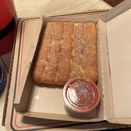 a box of bread and a container of ketchup - A rectangular garlic bread loaf topped with herbs and salt, served with a cup of marinara sauce in a pizza box.