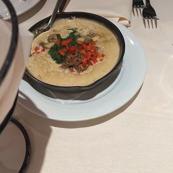 Roaring Fork - A bowl of creamy, possibly cheese or egg-based soup, garnished with chopped tomatoes, herbs, and small pieces of meat, placed on a white plate on a dining table. A person is holding a piece of bread or chip, and there are utensils, a napkin, and a glass of beverage nearby. - American Near Me - Austin, Texas