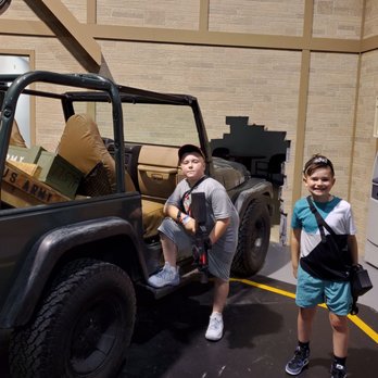 Tac Ops - It's A LOT darker during the game.... They put the lights on for the pictures afterwards. - Two boys are standing inside a building next to a military-style jeep with a cardboard box in the back. One boy is sitting on the vehicle's step, while the other stands beside it, both wearing casual clothing and carrying small bags. The background features a brick wall, an ATM machine, and a wall clock. The setting appears to be an indoor exhibit or museum. - Laser Tag Near Me - Fairfield, New Jersey