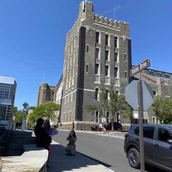 Cornell University - A historic stone building with a castle-like appearance stands at the corner of Stalter Drive, with pedestrians and cars nearby on a sunny day. - Colleges & Universities Near Me - Ithaca, New York