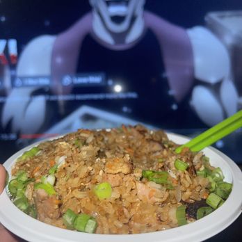Mampuku Ramen - Chicken fried rice - A bowl of fried rice with green onions is held in front of a TV screen displaying a character with a wide, open-mouthed smile and dark makeup. - Ramen Near Me - Santa Fe, New Mexico