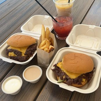 Twans Burger - Smashburger and Fries - Two cheeseburgers with beef patties, melted cheese, and buns are placed in takeout containers on a wooden table. A small paper container of French fries and two small cups of sauces are also present. Two drinks, one yellow and one red, with straws, are positioned behind the food. The setting appears to be outdoors, with a truck and some outdoor furniture visible in the background. - Food Trucks Near Me - Denver, Colorado