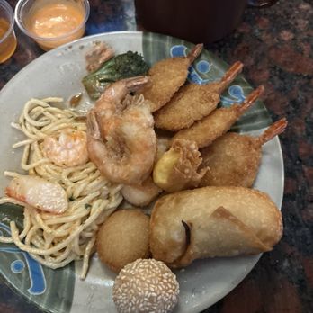 Sakura Buffet - Seafood Alfredo, Shrimp, Eggroll, sesame ball, Broccoli - A plate contains fried shrimp, a spring roll, sesame ball, fried potato, and fried cheese ball, alongside a serving of pasta with shrimp and broccoli. In the background, there is a cup of chips and small containers of dipping sauces. The food appears to be part of a buffet meal, with a dark, speckled countertop surface. - Sushi Bars Near Me - Jacksonville, Florida