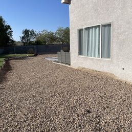 a green wagon parked in front of a house - An outdoor backyard space features a gravel ground surface adjacent to a white stucco house wall with a large window covered by curtains. To the right, a small green wagon is visible, and in the background, there is a wooden border separating the gravel from a grassy area. The scene is illuminated by sunlight, with trees and a clear blue sky in the distance.